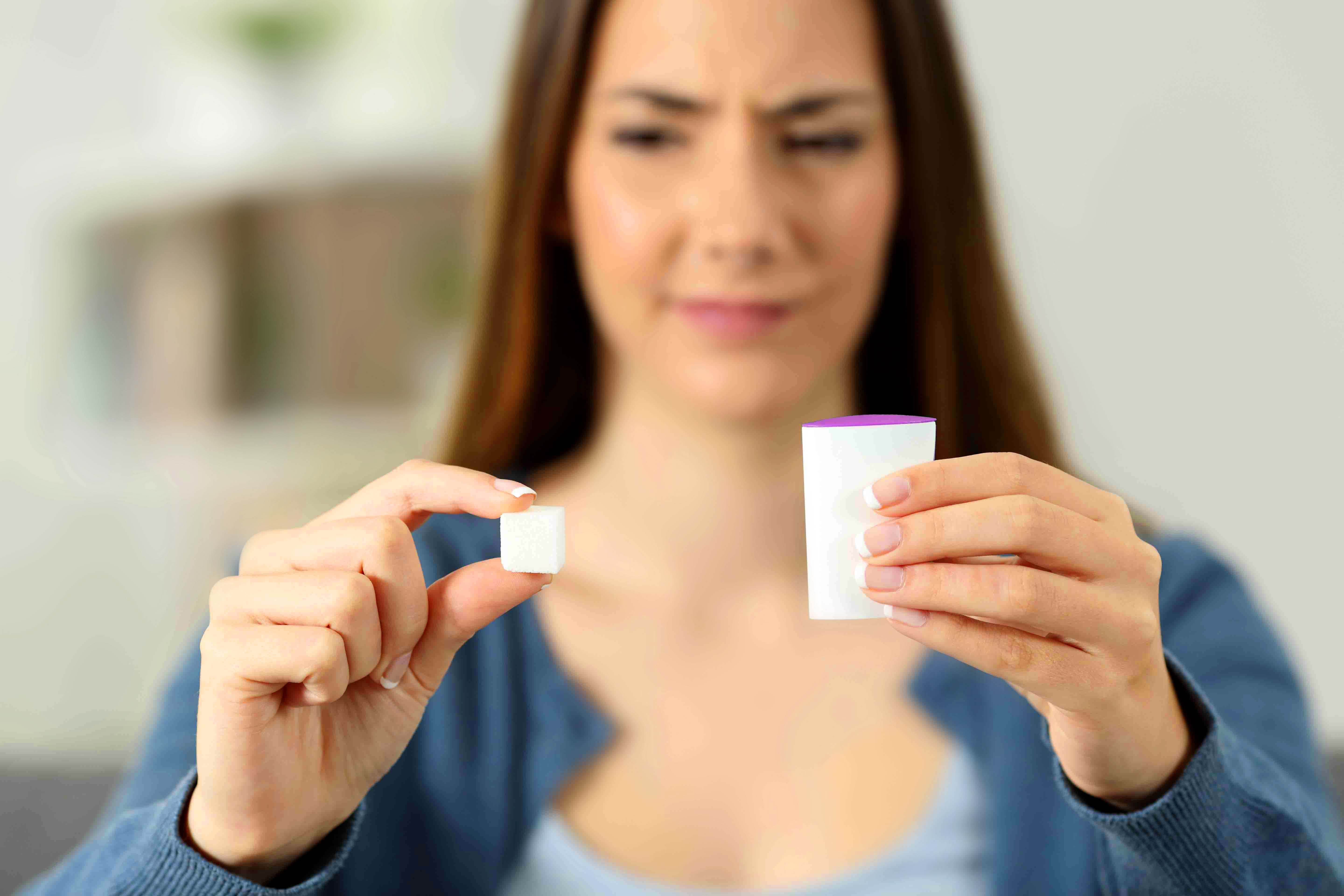 Leitartikel_Süßstoffe_Frau_und_Süßstoffe_verkleinert.jpg Eine Frau ,die skeptisch auf ein Stück Zucker und Süßstoff schaut