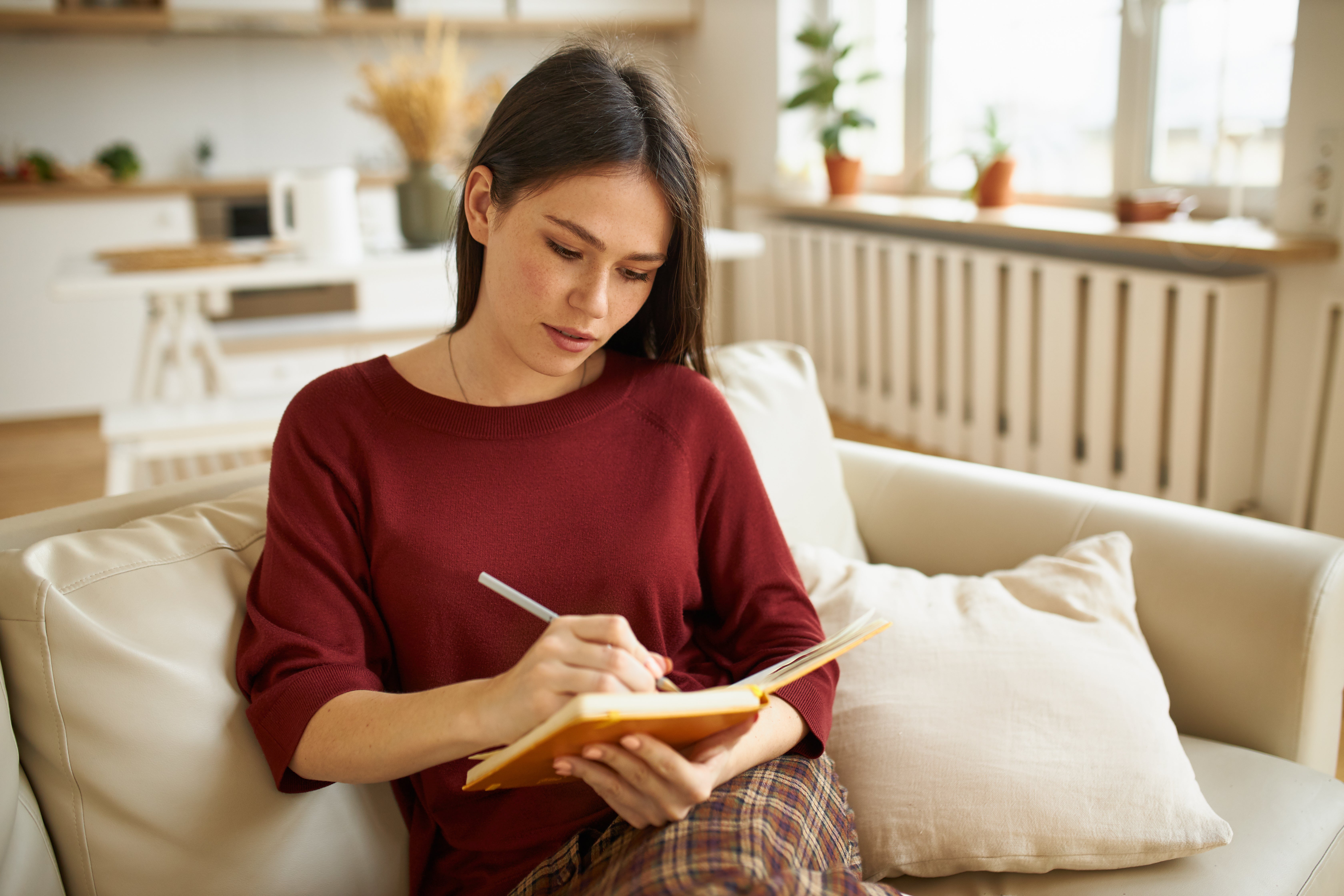 Frau sitzt auf Sofa und schaut in ihr Notizbuch rein, dabei hält sie den Stift in der rechten Hand.