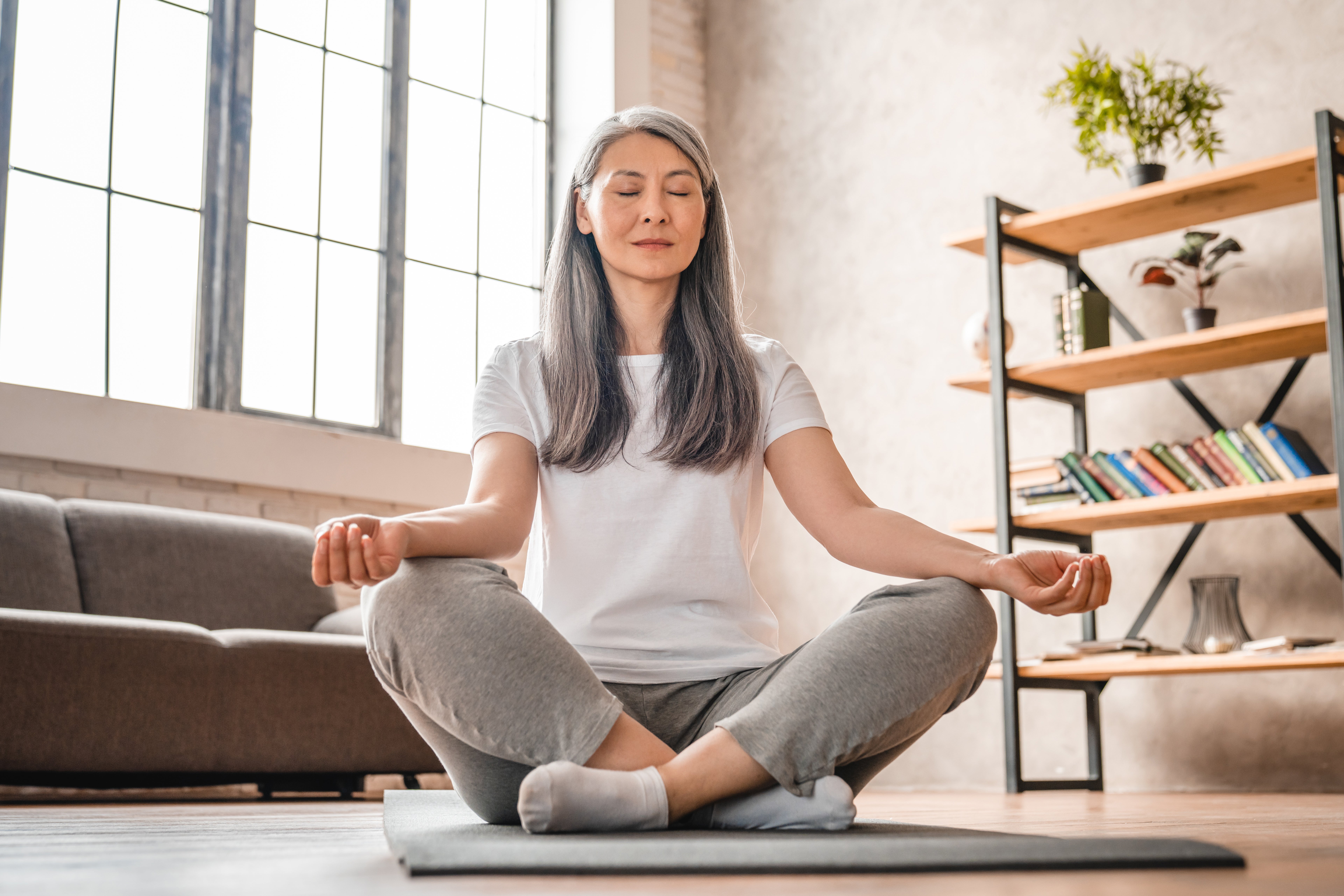 Frau sitzt im Yoga Schneidersitzt auf Boden, hinter ihr steht ein graues Sofa, sie hat die Augen geschlossen.