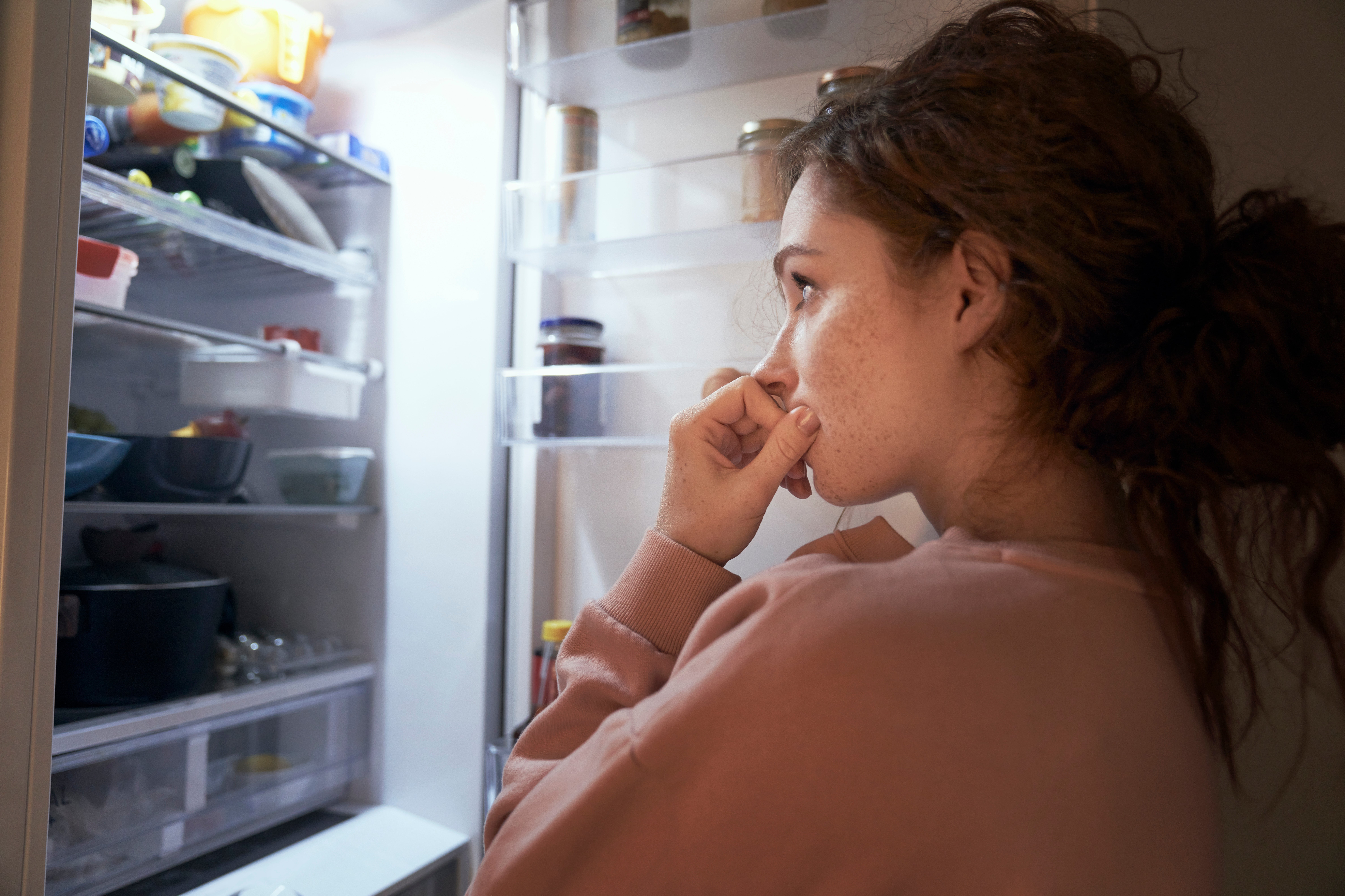 Frau_überlegt Man sieht eine Frau von der linken Seite wie sie vor einem offenen Kühlschrank steht und überlegt, was sie essen soll. "Appetitzügler gegen den Hunger?“
