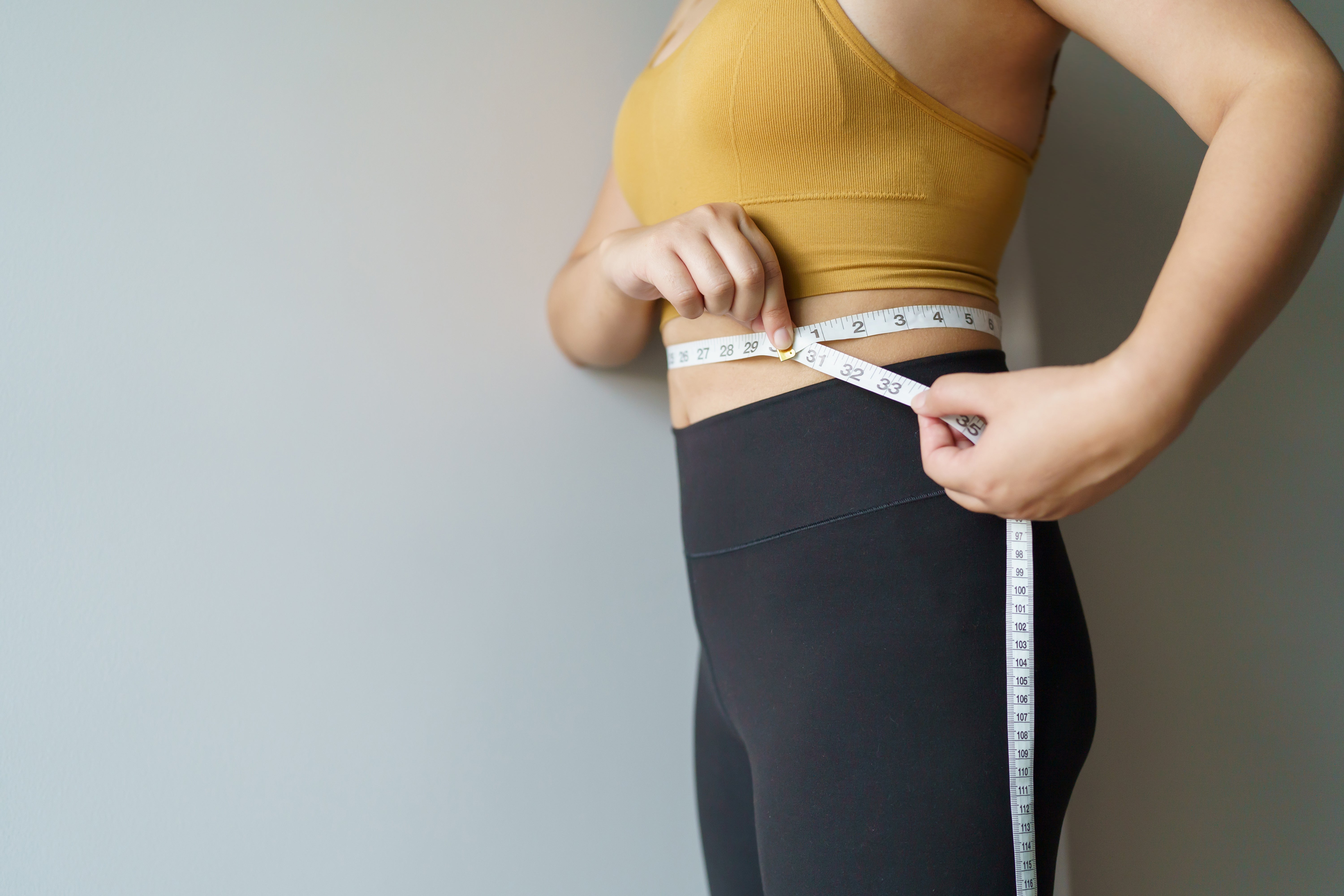 Frau_Bauchumfang Eine Frau, die mit einem Maßband ihren Bauchumfang misst. Sie trägt ein gelben Sport-BH und schwarze Leggins. Ihr Gesicht kann man nicht erkennen.