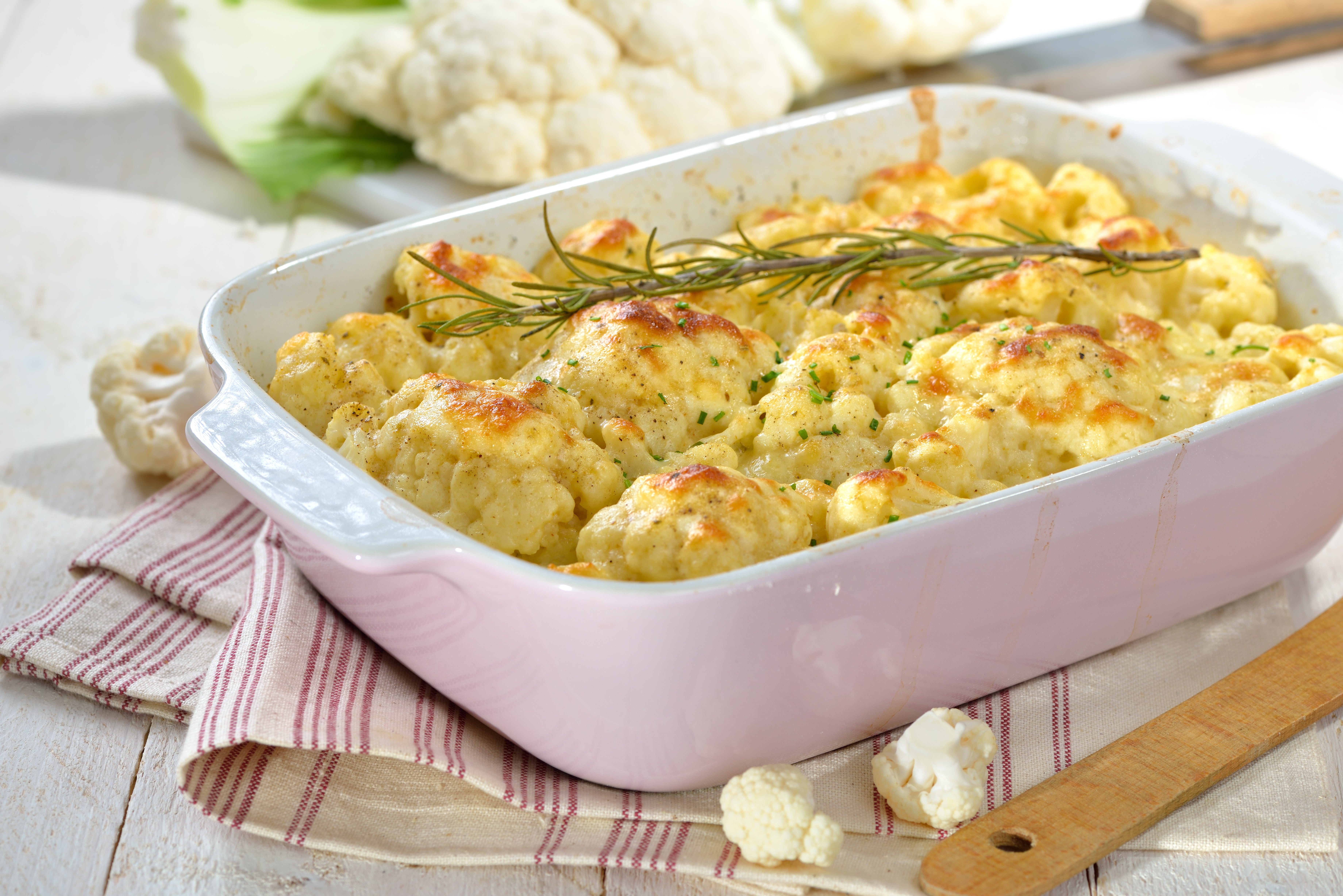 Käse überbacken Köstlich aussehender mit Käse überbackener Blumenkohl Auflauf in einer weißen Auflaufform auf einem rot weiß gestreiften Tuch. Auflauf ist mit Rosmarin dekoriert.