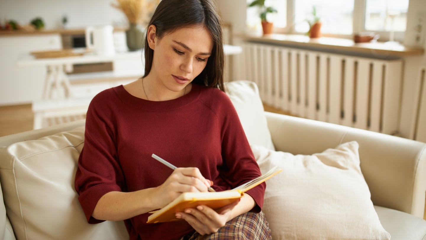 Frau sitzt auf Sofa und schaut in ihr Notizbuch rein, dabei hält sie den Stift in der rechten Hand.