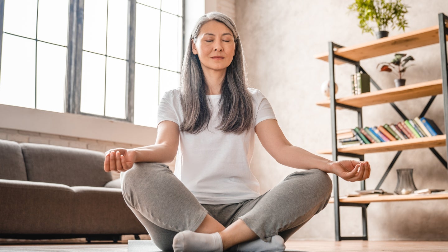 Frau sitzt im Yoga Schneidersitzt auf Boden, hinter ihr steht ein graues Sofa, sie hat die Augen geschlossen.
