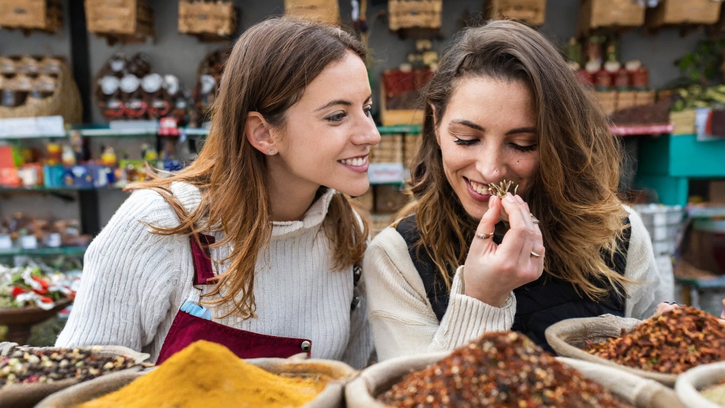 Zwei Frauen riechen an Gewürzen auf einem Gewürzmarkt. Beide Frauen lächeln.