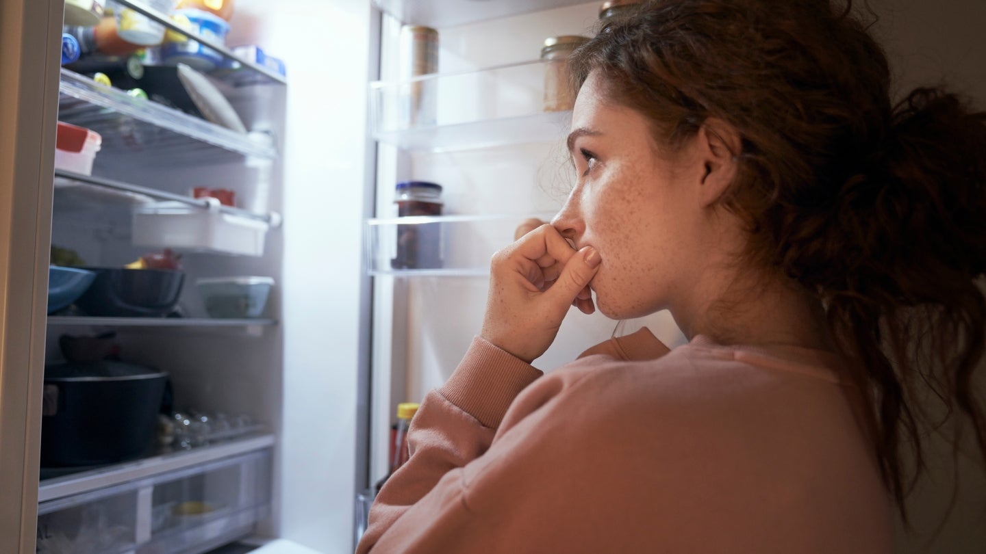 Frau_überlegt Man sieht eine Frau von der linken Seite wie sie vor einem offenen Kühlschrank steht und überlegt, was sie essen soll. "Appetitzügler gegen den Hunger?“