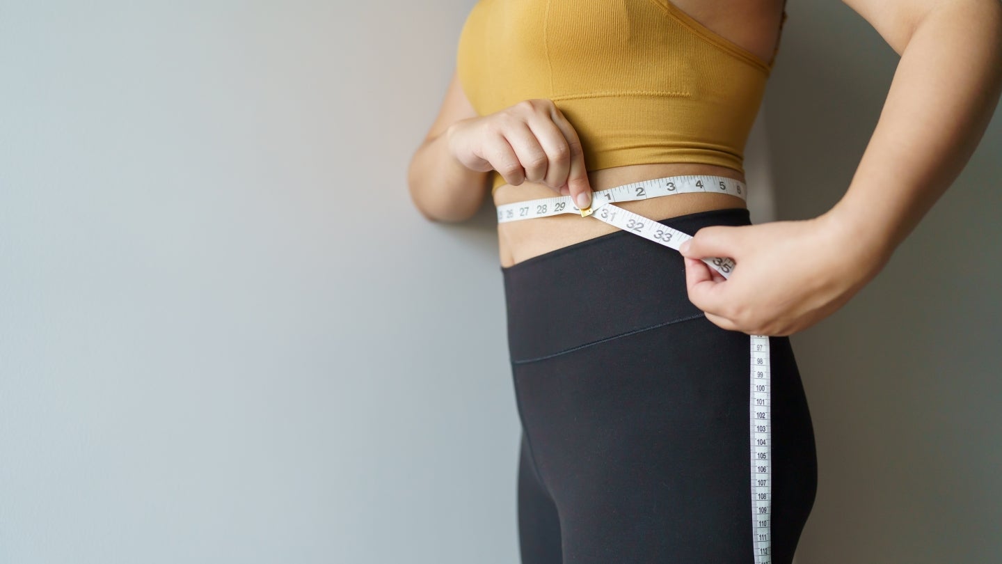 Frau_Bauchumfang Eine Frau, die mit einem Maßband ihren Bauchumfang misst. Sie trägt ein gelben Sport-BH und schwarze Leggins. Ihr Gesicht kann man nicht erkennen.