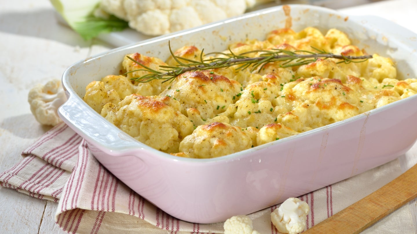 Käse überbacken Köstlich aussehender mit Käse überbackener Blumenkohl Auflauf in einer weißen Auflaufform auf einem rot weiß gestreiften Tuch. Auflauf ist mit Rosmarin dekoriert.