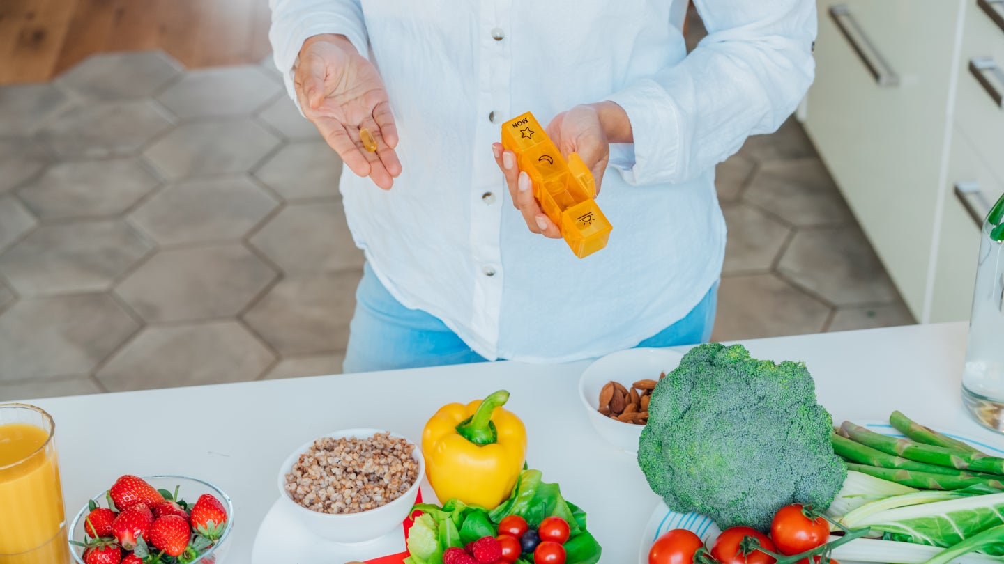 Frau hält Medikamenteorganisator, vor ihr liegen diverses Obst und Gemüse