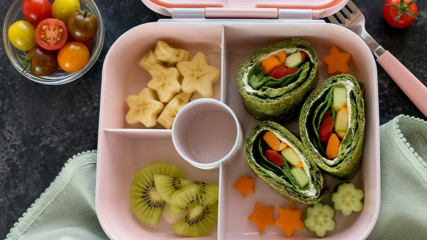 Spinat-Crepes in einer Lunchbox. Dazu Bananen und Kiwi und in einem kleinen Schälchen daneben kleine Tomaten.