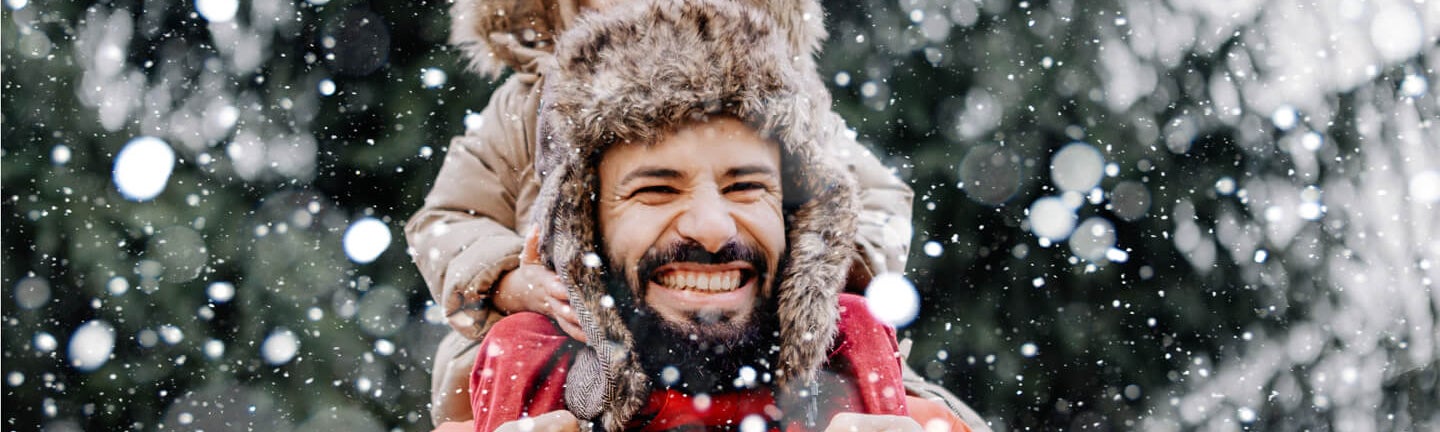 Ein Mann in Winterjacke trägt auf seinen Schulter ein kleines Kind draußen im Wald. Es schneit und der Mann lächelt glücklich