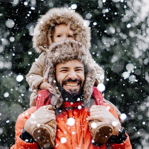 Ein Mann in Winterjacke trägt auf seinen Schulter ein kleines Kind draußen im Wald. Es schneit und der Mann lächelt glücklich