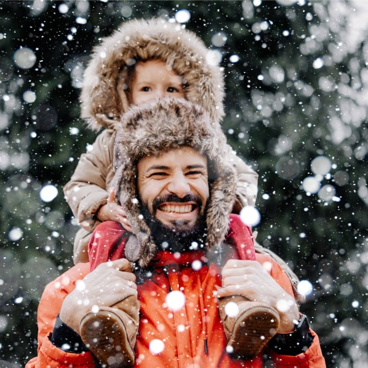 Ein Mann in Winterjacke trägt auf seinen Schulter ein kleines Kind draußen im Wald. Es schneit und der Mann lächelt glücklich