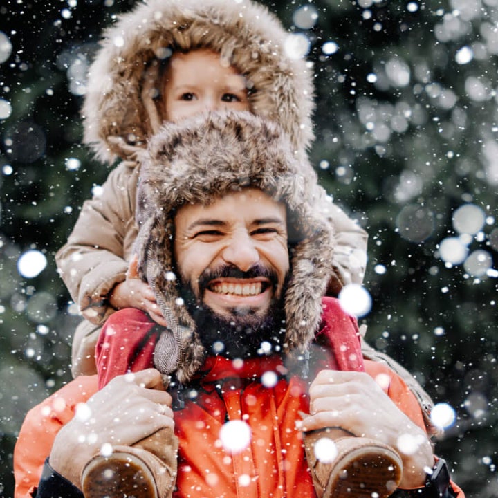 Ein Mann in Winterjacke trägt auf seinen Schulter ein kleines Kind draußen im Wald. Es schneit und der Mann lächelt glücklich